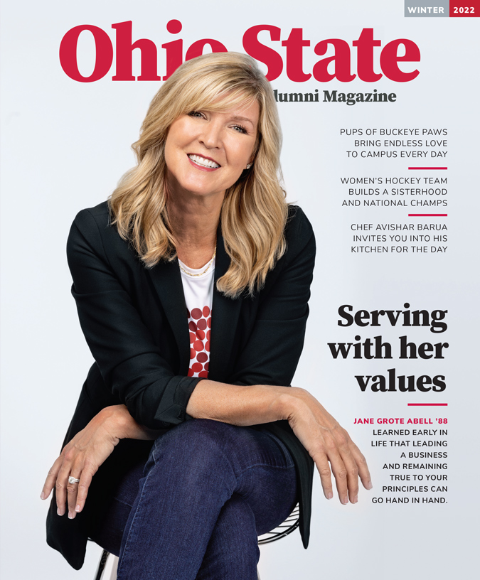 A white lady with long blond hair smiles like she's greeting a friend while wearing a pizza T-shirt and dark blazer. She's sitting on a stool and slightly leaning forward.