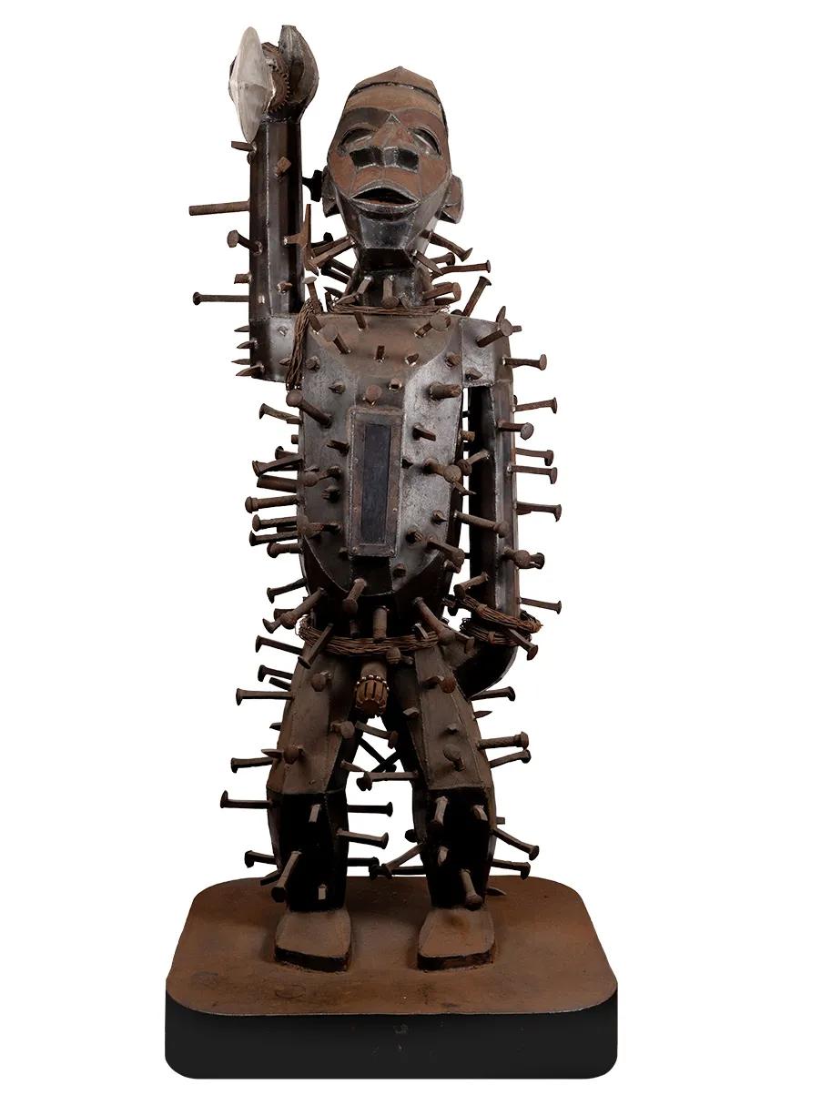 A metal statue with a weathered patina shows an African warrior holding up his sword with thick nails or spikes hammered all over his body. The exception is his face—none is higher than his chin. The warrior looks straight forward. What would usually be rounded—shoulders, belly, arc of the cheek, is rendered in flat panels of metal, adding a geometric look to the warrior.
