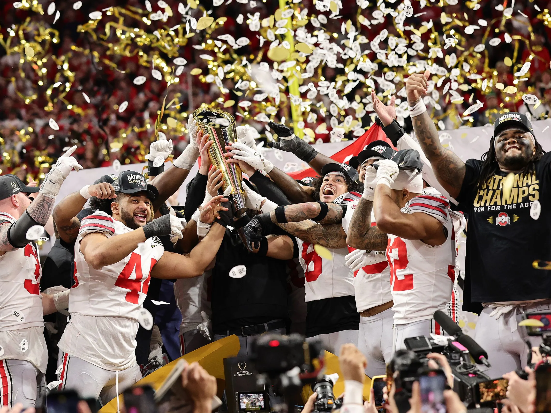 Buckeye football players cheer as they hoist up the national championship trophy. Confetti falls and they're amidst a huge crowd.