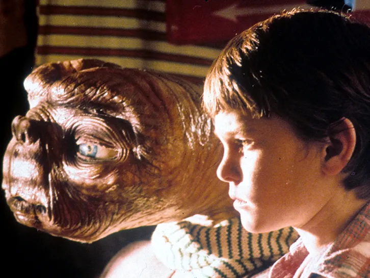 In a still from the movie ET, the alien and a little boy look out a window in a dim room.