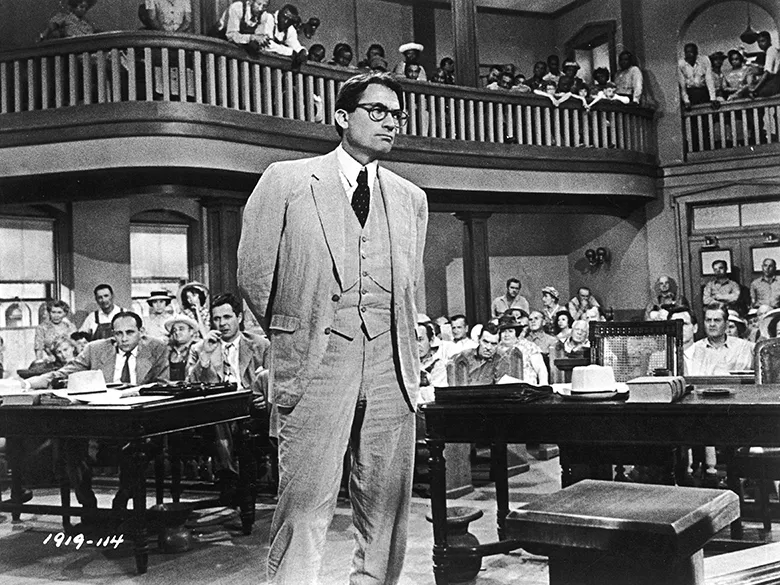 In this photo from an old movie, a white man in a suit stands in the middle of a courtroom