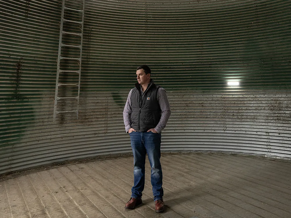 Will Hamman, a young white man wearing jeans and an insulated vest over his plaid button-down, stands inside an empty grain silo on his family farm in Ohio. The structure is corrugated metal maybe five times as wide as he is tall, with a peaked roof. A ladder and big flood light are too his right.