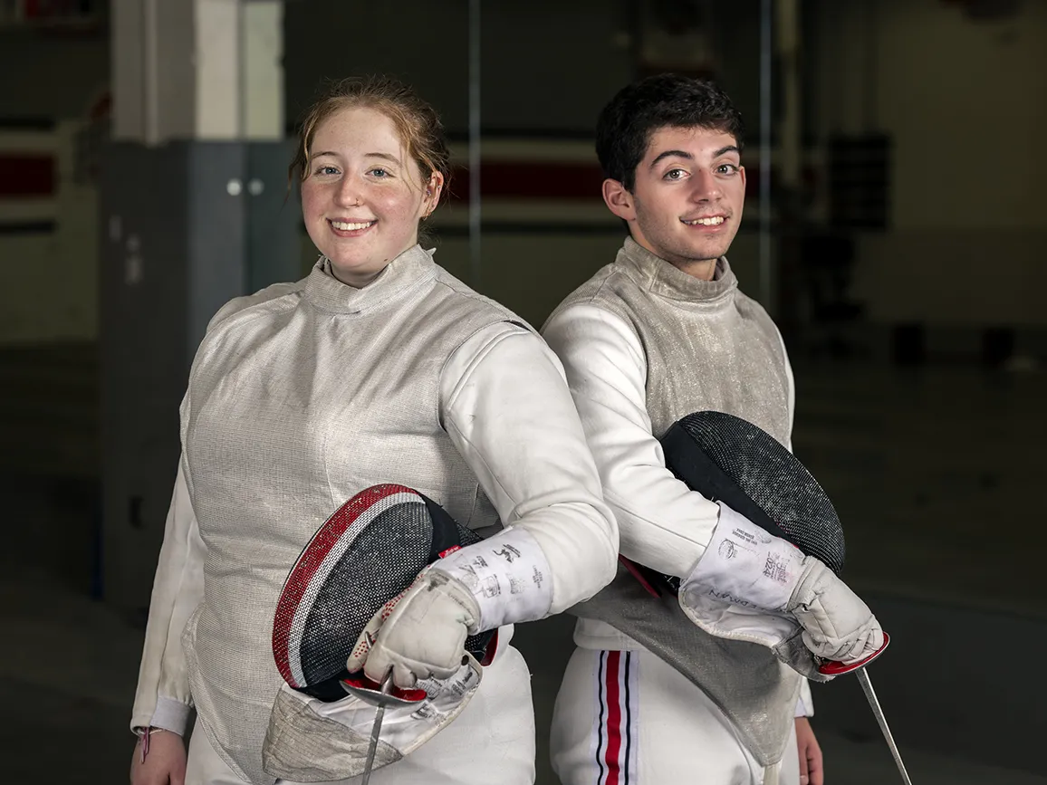 Siblings Miranda and Sammy Freedman smile as they pose for a photo in their fencing uniforms. Both are gray with use--they look like they've enjoyed a lot of competition time in them.