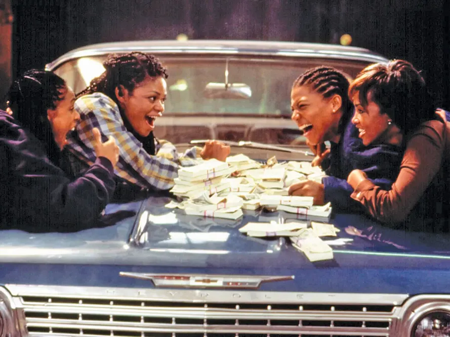 In this still from a movie, four young black women lean over the hood of a car with cash piled in the middle. They're laughing joyfully.