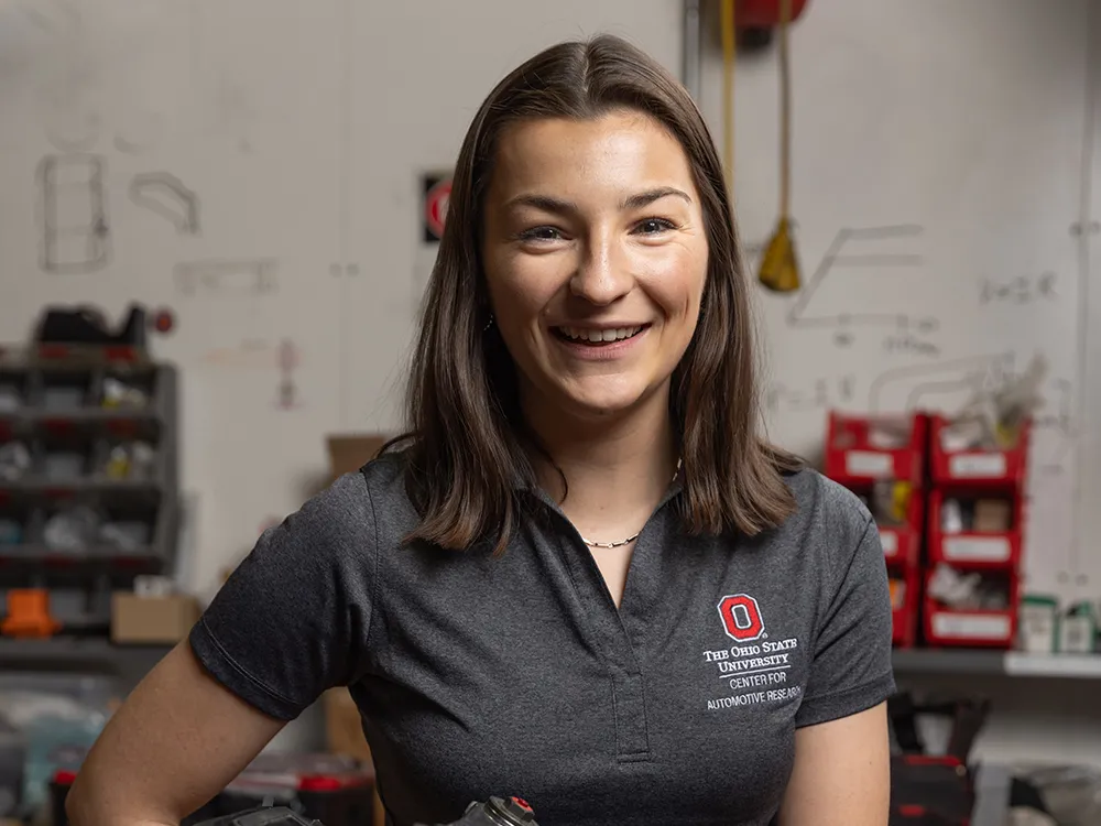 Laura Friedmann wears an Ohio State polo shirt branded Center for Automotive Research, as she smiles in this portrait in a motorcycle laboratory. She is a young woman with shoulder-length hair and her smile says she is genuinely thrilled to be photographed.